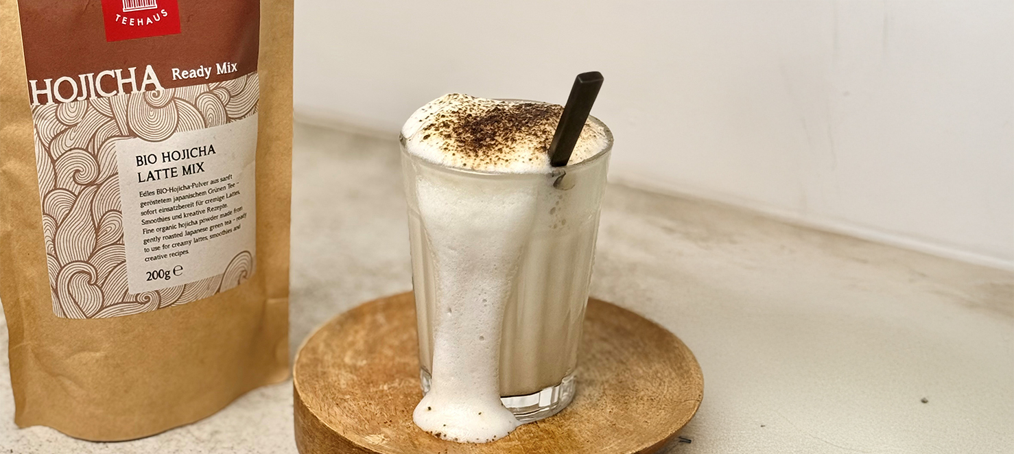 Bio Hojicha Latte Mix von Demmers Teehaus neben Glas Hojicha Latte mit Milchschaum und Hojicha-Pulver auf Holzuntersetzer