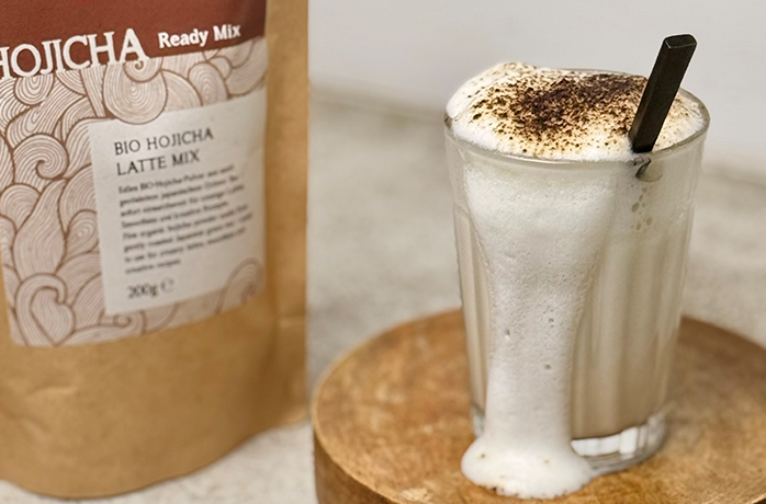 Bio Hojicha Latte Mix von Demmers Teehaus neben Glas Hojicha Latte mit Milchschaum und Hojicha-Pulver auf Holzuntersetzer