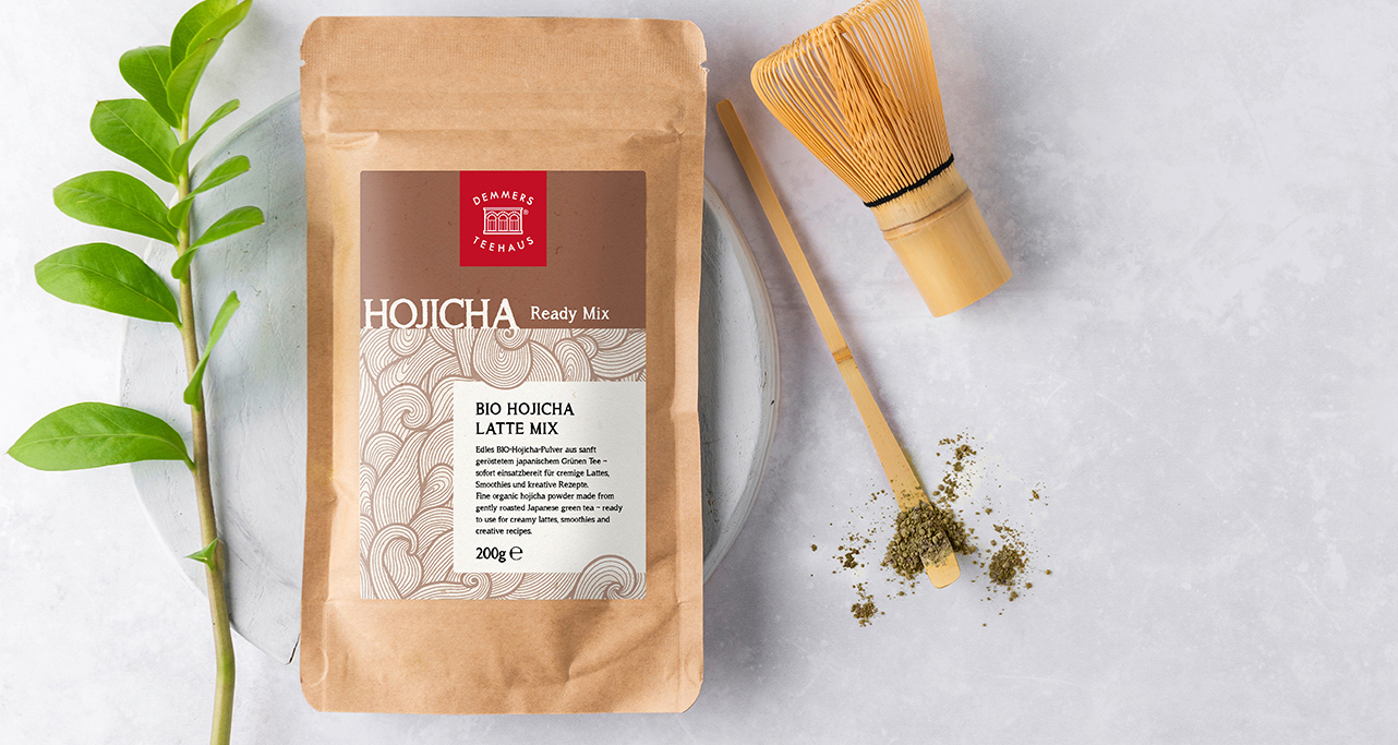 Demmers Teehaus Bio Hojicha Latte Mix Ready Mix (200 g) in Papierverpackung mit Bambusbesen und Hojicha-Pulver auf hellem Untergrund
