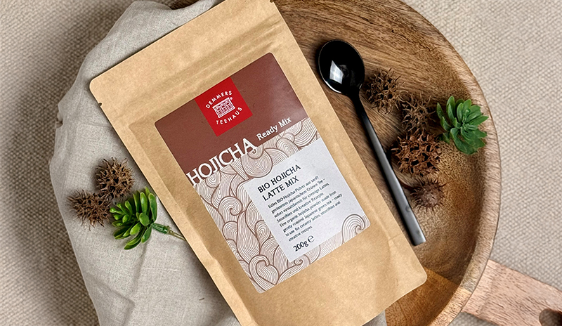 Bio Hojicha Latte Mix Ready Mix von Demmers Teehaus (200 g) auf Holzbrett mit schwarzem Löffel und grüner Pflanze, Draufsicht