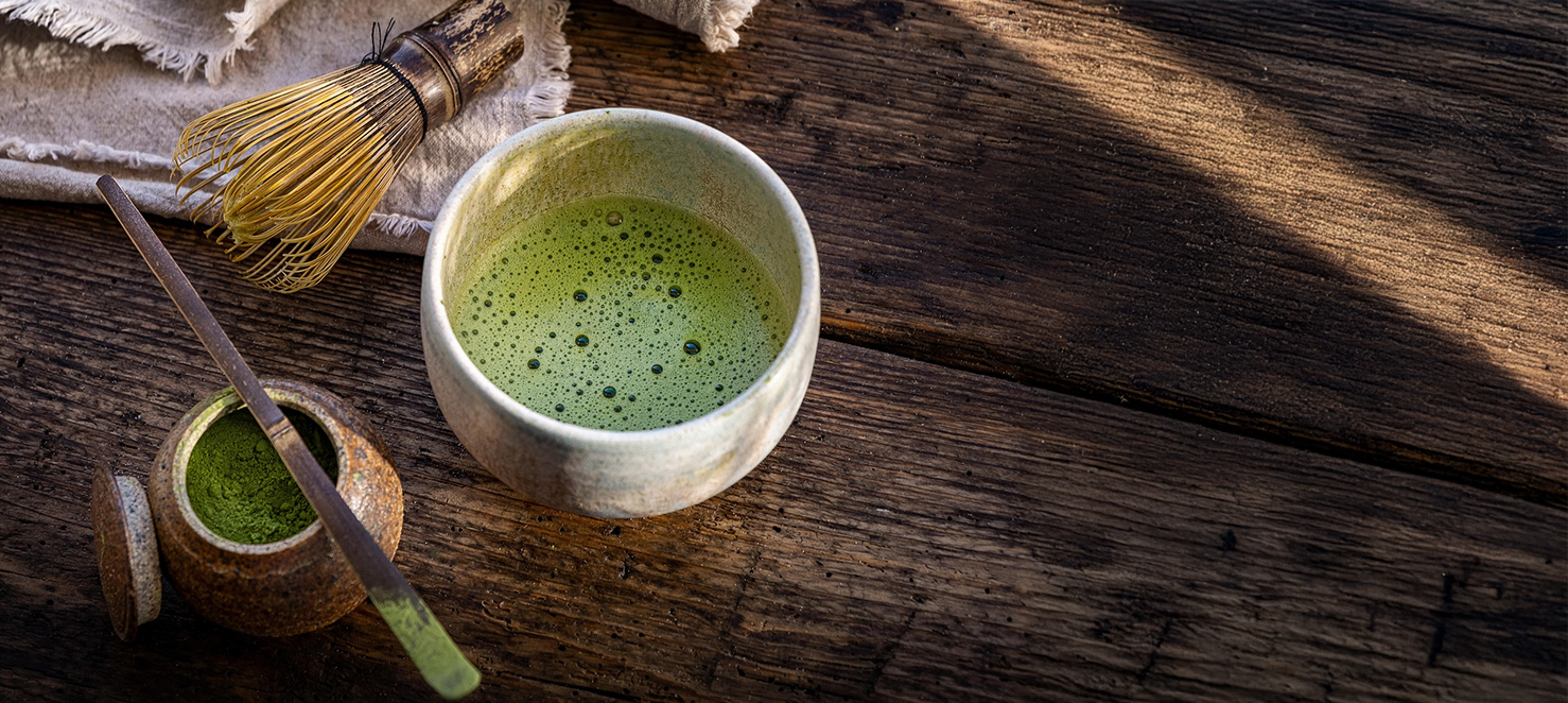Frisch zubereiteter Matcha-Tee in einer Keramikschale mit Matcha-Pulver im Döschen, Bambusbesen und Leinentuch auf einem Holztisch