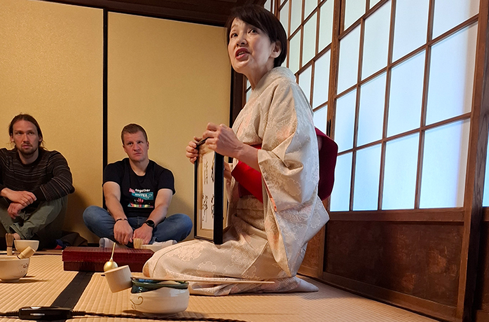 Japanische Teemeisterin im Kimono erklärt im traditionellen Tatami-Raum einer Besuchergruppe die Teezeremonie