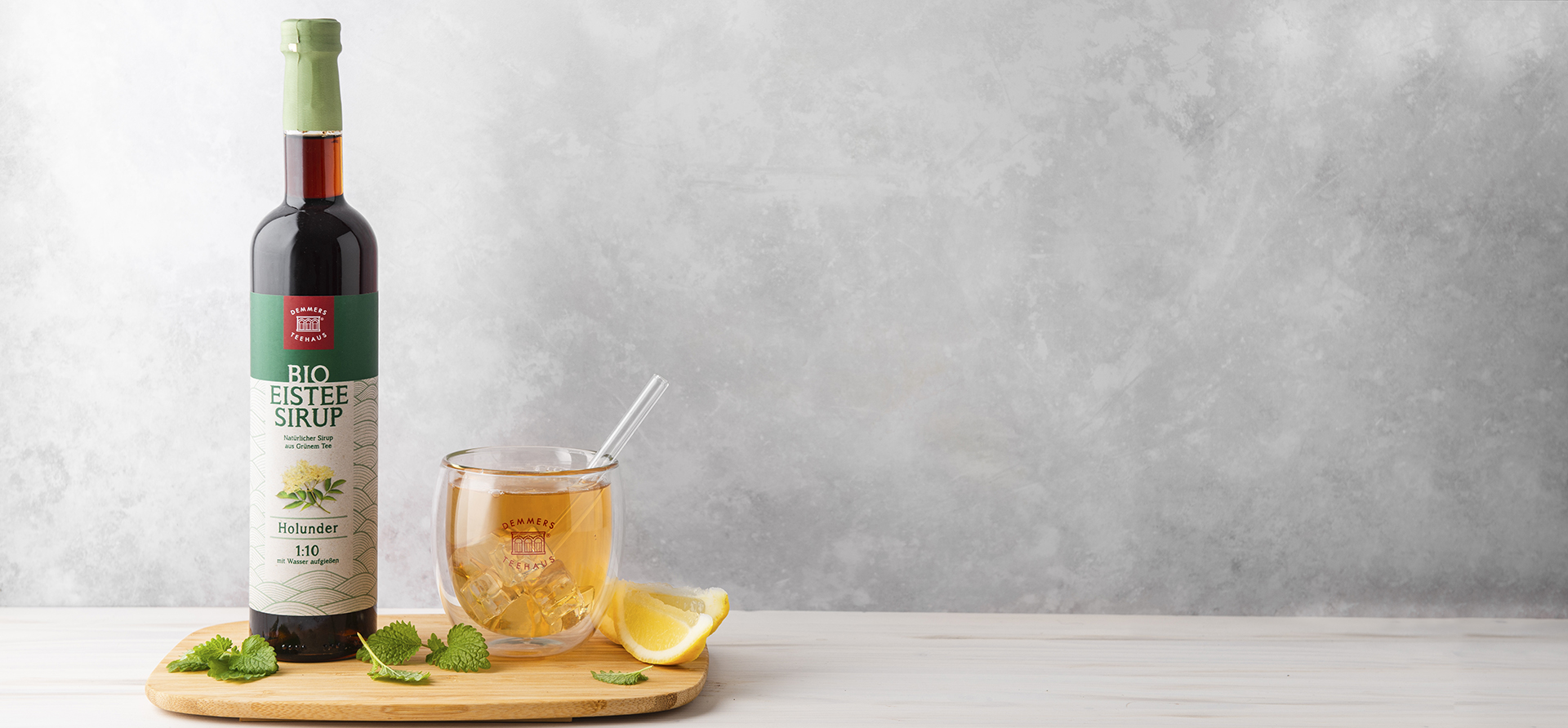 Demmers Teehaus BIO Eistee-Sirup Grüner Tee Holunder Flasche neben Glas mit kaltem Eistee und Limetten auf Holztablett 