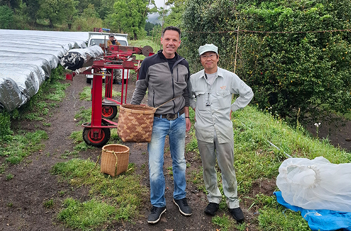 Martin gemeinsam mit einem Mitarbeiter der Teeplantage in Japan – mit Pflückkorb und Erntemaschinen im Hintergrund Martin gemeinsam mit einem Mitarbeiter der Teeplantage in Japan – mit Pflückkorb und Erntemaschinen im Hintergrund