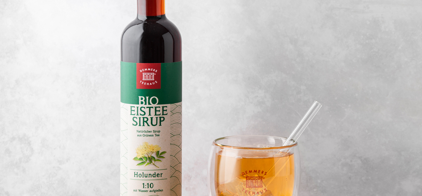 Demmers Teehaus BIO Eistee-Sirup Grüner Tee Holunder Flasche neben Glas mit kaltem Eistee und Limetten auf Holztablett
