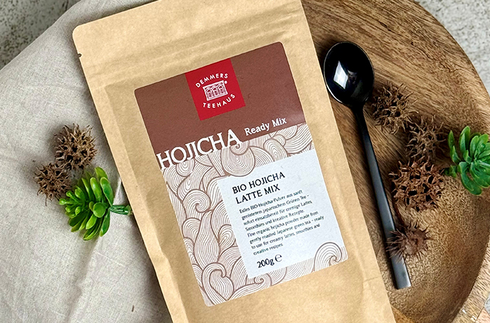 Bio Hojicha Latte Mix Ready Mix von Demmers Teehaus (200 g) auf Holzbrett mit schwarzem Löffel und grüner Pflanze, Draufsicht